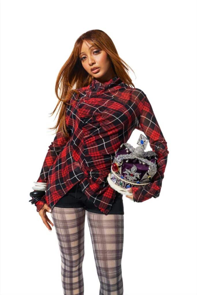 Young woman in a red and black plaid top, plaid tights and black shorts poses against a white background while holding an ornate jeweled crown. Promotional portrait for a music festival.