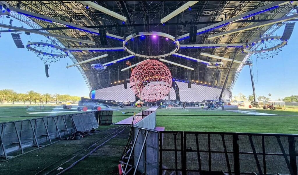 Large open-air concert stage under a curved canopy with a suspended geodesic spherical chandelier and radial lighting rig; crew and equipment on artificial turf prepare the stage while metal crowd barricades run across the foreground.