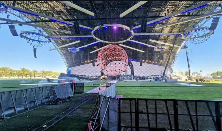 Large open-air concert stage under a curved canopy with a suspended geodesic spherical chandelier and radial lighting rig; crew and equipment on artificial turf prepare the stage while metal crowd barricades run across the foreground.