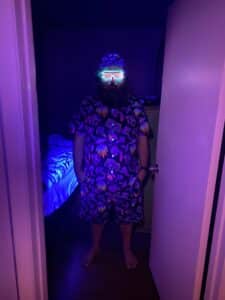 Person stands barefoot in a doorway wearing a matching UV‑reactive mushroom‑print shirt, shorts and bucket hat, with glowing neon shutter shades under purple and blue lighting. The outfit and lighting create a festival‑ready, blacklight‑reactive look.