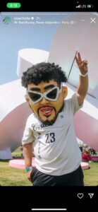 Person wearing an oversized caricature Bad Bunny mask with curly hair, thick eyebrows and white sunglasses flashes a peace sign while standing in front of a large pink-and-white sculpture at an outdoor festival; they wear a white shirt marked "23" and multiple wristbands.