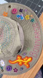 Handmade khaki fisherman’s hat decorated with concentric rainbow running stitches on the crown, embroidered flowers and the word Bonnaroo stitched along the brim, plus peace-sign buttons, a small bee patch, an orange lizard appliqué and scattered beads. A needle and thread are visible, indicating it’s being prepared for festival wear.