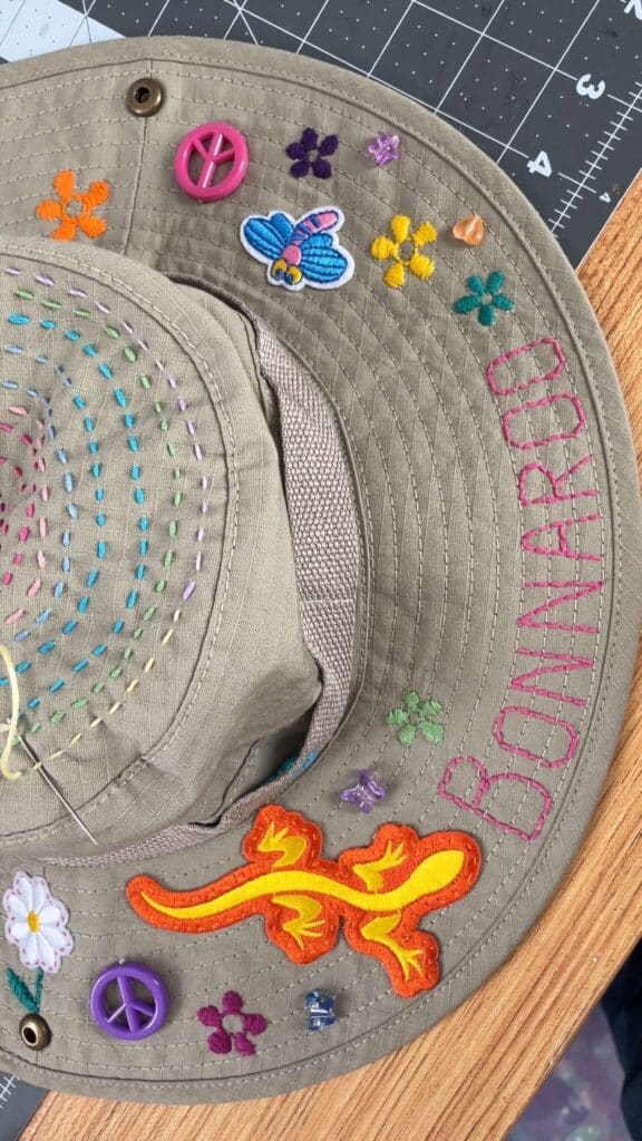 Handmade khaki fisherman’s hat decorated with concentric rainbow running stitches on the crown, embroidered flowers and the word Bonnaroo stitched along the brim, plus peace-sign buttons, a small bee patch, an orange lizard appliqué and scattered beads. A needle and thread are visible, indicating it’s being prepared for festival wear.