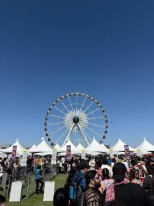 Large Ferris wheel towers over a packed festival entrance where white tents and “scan point” signs mark multiple security lanes as long lines of attendees wait to be scanned under a clear blue sky.