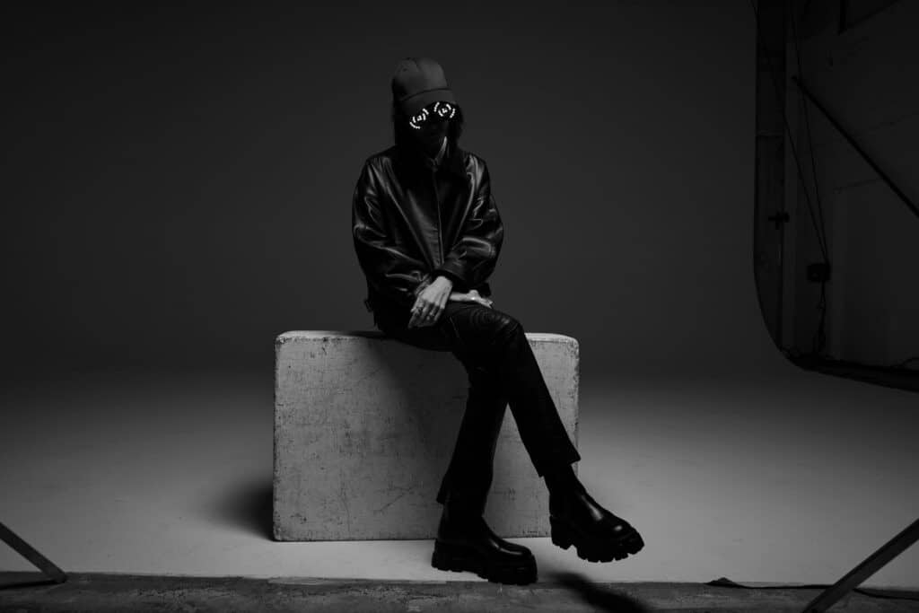 A performer sits on a rectangular studio block in moody black-and-white lighting, wearing a leather jacket, baseball cap, and LED goggles that glow with concentric circular patterns, face mostly obscured. They cross their legs and wear chunky platform boots, creating a dark, futuristic DJ aesthetic.