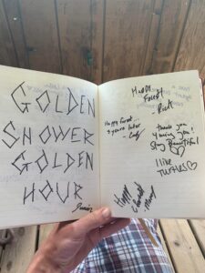 Open notebook held by a person's hand shows two pages filled with handwritten messages and signatures; the left page has large blocky letters reading "GOLDEN SHOWER GOLDEN HOUR" and a small "Junior" signature. The right page contains assorted notes like "Happy Forest!", "thank you 4 being you! Stay Beautiful", and scribbled names, with a wooden deck and plaid shorts visible in the background.