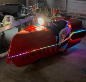 Custom white and red motor scooter fitted with oversized red side pods trimmed with fringe and multicolored LED strips, headlight on and parked in a garage workshop.