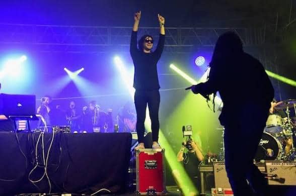 Performer in black clothing and sunglasses stands on a red road case at center stage with arms raised while colorful purple and green stage lights illuminate DJ equipment, band members, and a photographer crouching nearby.