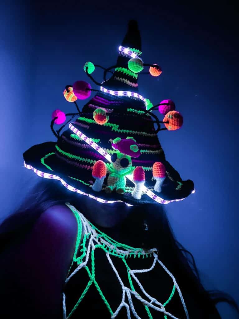 Person wearing a tall crochet witch hat wrapped with purple LED strip lights and decorated with neon pom-poms, a tiny crocheted alien figure and small mushrooms. A glowing crochet net top and deep blue background emphasize the blacklight-reactive colors.
