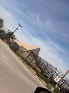 Beige triangular temporary structure resembling a pyramid sits behind a tall metal security fence topped with pointed bars at a festival build site, with a "No Lacrosse Access" sign visible and palm trees against a blue sky in the background.