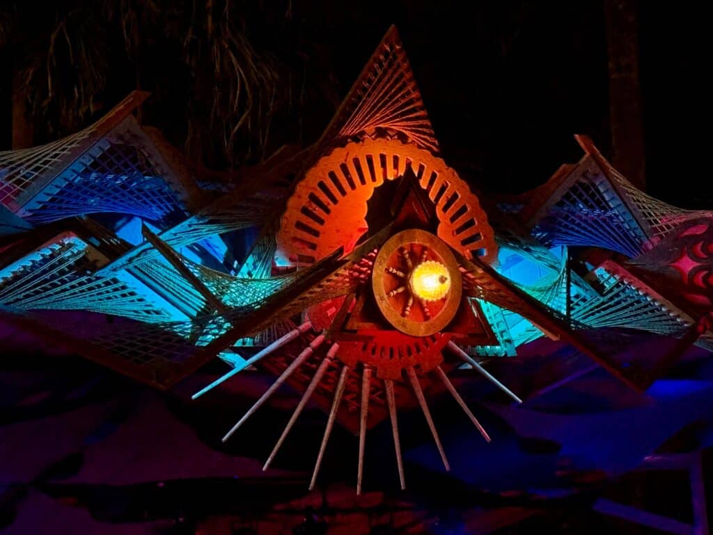 Illuminated geometric wooden sculpture suspended overhead at night with a central amber bulb surrounded by radial spokes and gear-like cutouts, flanked by triangular lattice panels lit in teal and magenta.