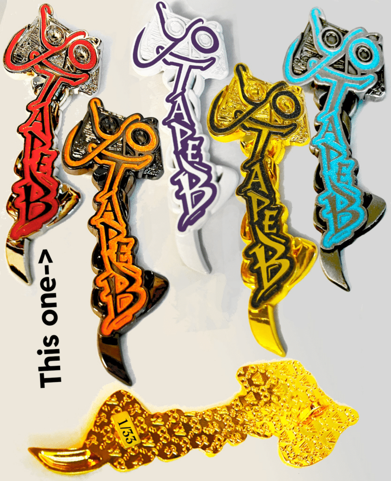 Colorful enamel pins shaped like a stylized key or dagger with graffiti-style "TABEB" lettering in red, orange, purple, gold, turquoise and black, arranged on a white background; a gold-backed pin shows a stamped "1/33" edition number. Bold black text and an arrow label one pin as "This one" to highlight the giveaway selection.