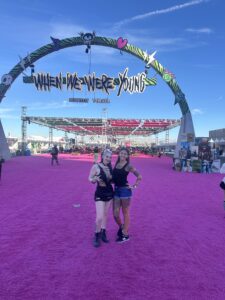 Two women in black outfits pose and smile on a bright magenta carpet under a large decorative arch that reads "When We Were Young," with a covered stage, booths, and festival attendees visible in the background beneath a clear blue sky.