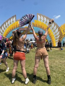 Two young women stand with their arms raised toward a large colorful festival arch labeled Electric Forest, wearing crop tops and short skirts while one holds a decorative fan, as a crowd gathers on the grassy field under a blue sky.
