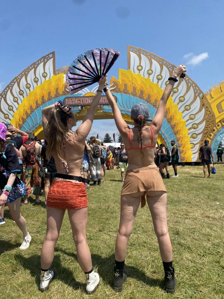 Two young women stand with their arms raised toward a large colorful festival arch labeled Electric Forest, wearing crop tops and short skirts while one holds a decorative fan, as a crowd gathers on the grassy field under a blue sky.