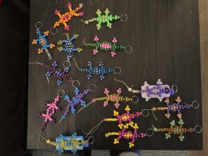 Several colorful beaded lizard keychains arranged on a dark tabletop, each made from plastic pony beads and attached to metal split rings in various color combinations. They appear handmade as DIY festival freebies, showing different patterns and sizes.