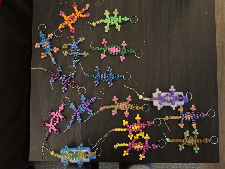 Several colorful beaded lizard keychains arranged on a dark tabletop, each made from plastic pony beads and attached to metal split rings in various color combinations. They appear handmade as DIY festival freebies, showing different patterns and sizes.