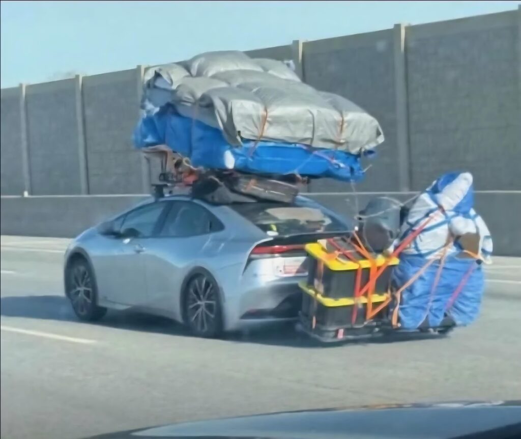 Silver hatchback on a highway carrying an enormous load—multiple blue and gray tarped bundles stacked on the roof and a bulky, strap-secured cargo rack extending from the rear filled with suitcases and camping gear.